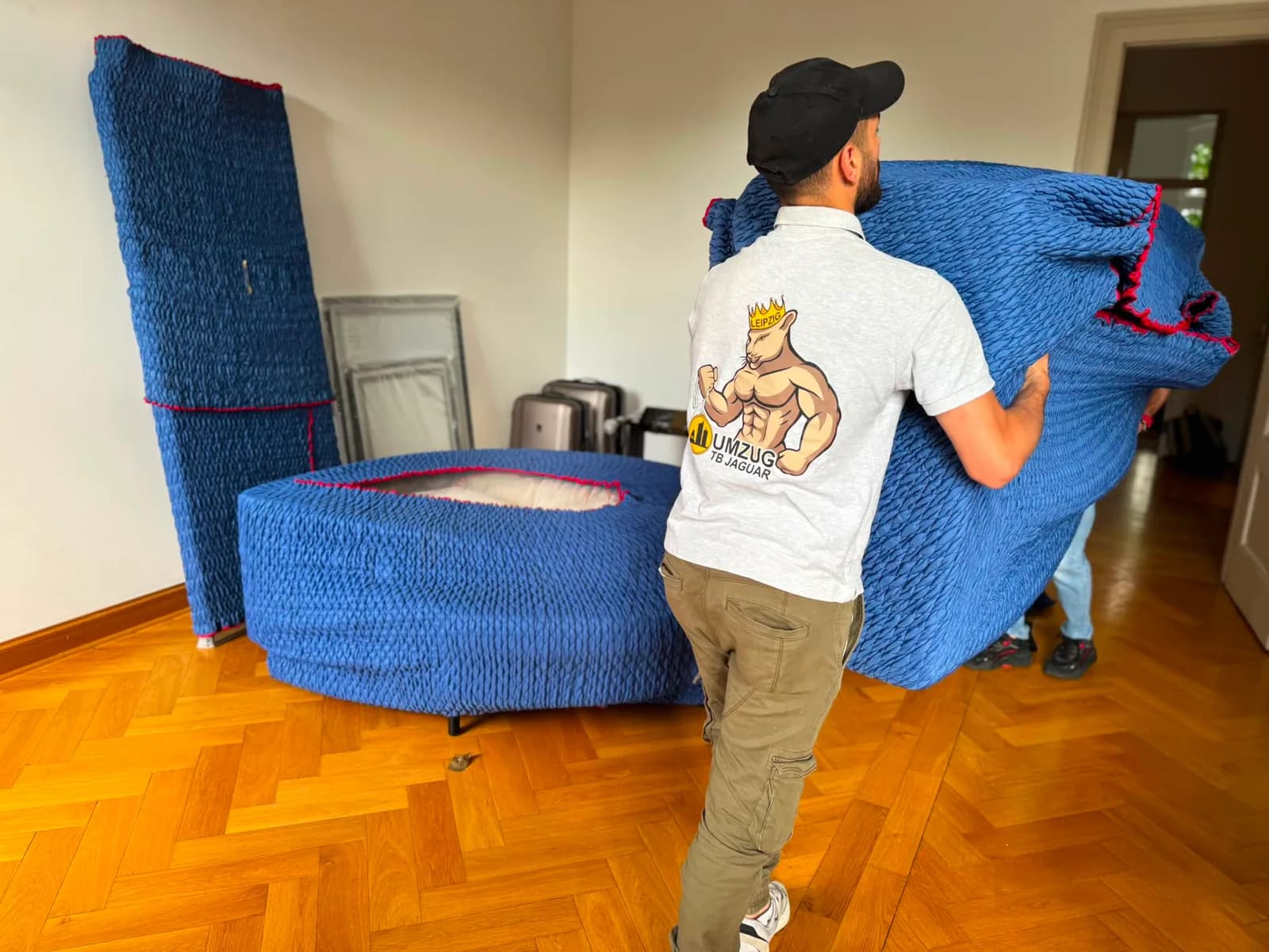 Umzugshelfer von TB Jaguar trägt ein in eine blaue Umzugsdecke eingeschlagenes Polstermöbel durch ein Schlafzimmer